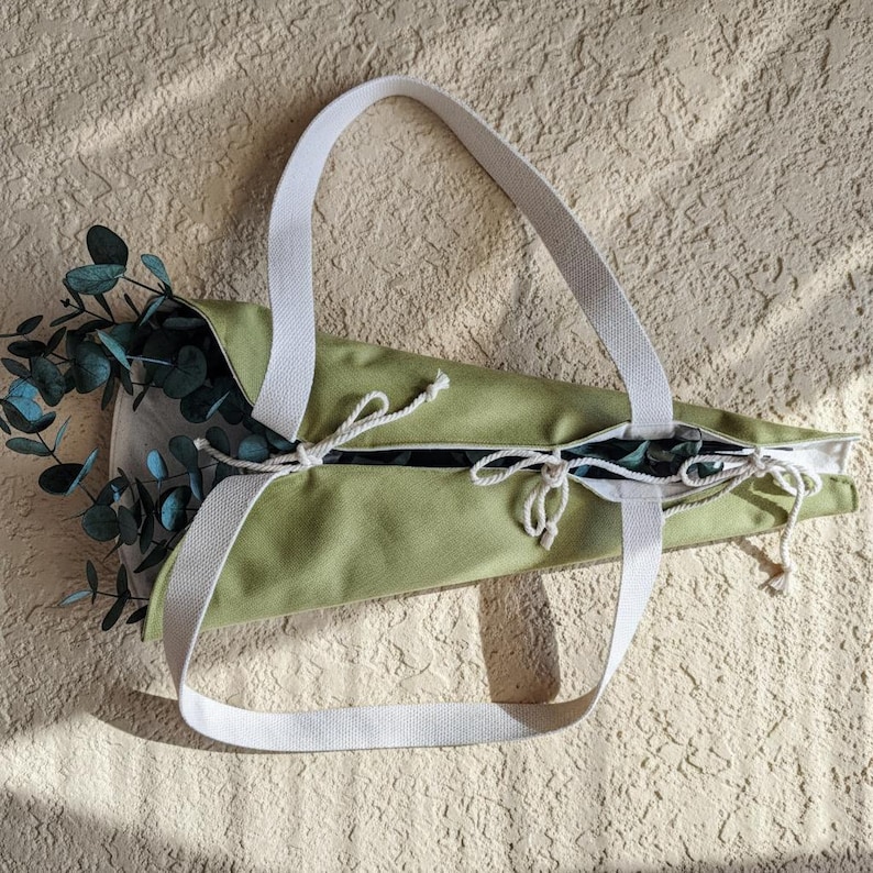 Puede incluir: Un bolso de tela de lona en color verde claro con asas de tela blanca y cordones blancos. El bolso tiene forma c&oacute;nica y contiene una rama de hojas de eucalipto de color verde oscuro que sobresale por la abertura superior.