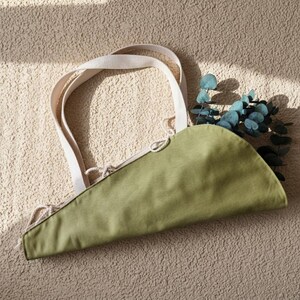Puede incluir: Un bolso de tela verde oliva en forma de semic&iacute;rculo con dos asas tejidas en color blanco roto y un cierre de cord&oacute;n con finas cuerdas. El bolso est&aacute; parcialmente relleno con una rama de hojas de eucalipto de color verde.