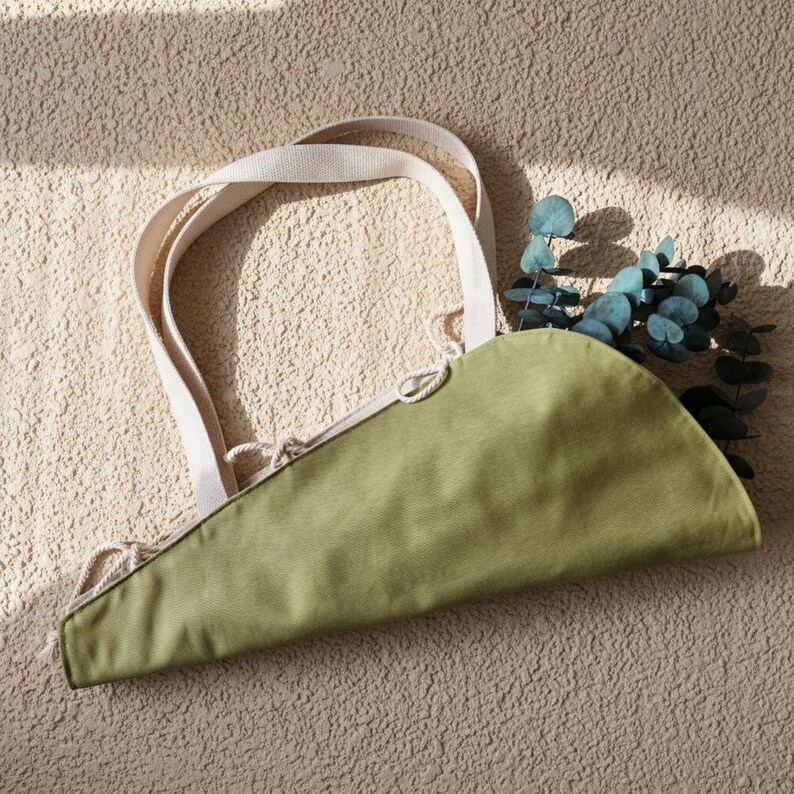 Puede incluir: Un bolso de tela verde oliva en forma de semic&iacute;rculo con dos asas tejidas en color blanco roto y un cierre de cord&oacute;n con finas cuerdas. El bolso est&aacute; parcialmente relleno con una rama de hojas de eucalipto de color verde.