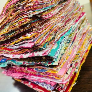 Könnte beinhalten: Ein Stapel bunter Stoffreste in verschiedenen Mustern und Farbtönen, darunter Rosa, Gelb, Grün und Rot. Die Kanten des Stoffes sind sichtbar und erzeugen ein strukturiertes Aussehen. Das Bild zeigt eine Sammlung von Textilresten, ideal für Bastel- oder Quiltprojekte.