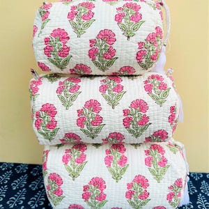 Könnte beinhalten: Drei weiße, gesteppte Kosmetiktaschen, die übereinander gestapelt sind. Jede Tasche ist mit einem sich wiederholenden Muster aus rosa Blumen und grünen Blättern verziert. Die Taschen haben eine rechteckige Form mit abgerundeten Kanten und einem Reißverschluss.