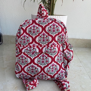 May include: A red and white floral patterned backpack with a quilted design. The backpack features a front pocket, adjustable straps, and a top handle. The floral pattern includes pink flowers and green leaves.