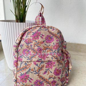 Puede incluir: Mochila acolchada con estampado floral en rosa, naranja y azul sobre fondo beige. La mochila tiene un bolsillo frontal y un asa superior. La bolsa mide aproximadamente 30 cm de alto y 25 cm de ancho.