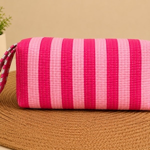 May include: A rectangular quilted pouch featuring vertical stripes in alternating shades of hot pink and light pink. The pouch includes a matching fabric wrist strap with a small white geometric pattern and a visible quilted texture throughout.