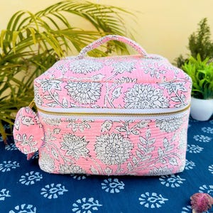 Peut inclure: Trousse de maquillage rose à imprimé floral blanc. Elle est dotée d'une fermeture éclair dorée, d'une poignée et d'un pompon assorti. Le sac est posé sur un tissu bleu à motifs. Dimensions : environ 20 cm x 15 cm x 15 cm.