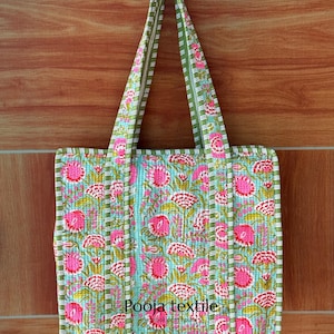 May include: A quilted tote bag with a floral pattern in pink and white on a light blue background. The bag has green and white striped handles and trim. The text "Pooja textile" is printed on the bottom of the bag.