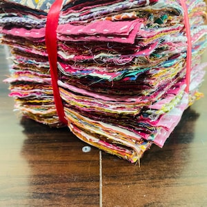 May include: A stack of colorful fabric squares, tightly bound with a red ribbon. The squares display a variety of patterns and colors, including pinks, blues, yellows, and floral designs. The edges of the fabric are visible, creating a textured appearance.