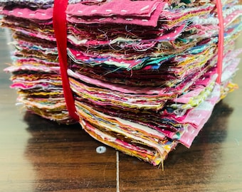 May include: A stack of colorful fabric squares, tightly bound with a red ribbon. The squares display a variety of patterns and colors, including pinks, blues, yellows, and floral designs. The edges of the fabric are visible, creating a textured appearance.