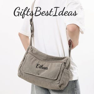 Puede incluir: Un bolso mensajero gris con el nombre "Ethan" bordado en la solapa delantera. El bolso tiene un bolsillo delantero, un bolsillo lateral y una correa para el hombro. El texto "Gifts Best Ideas" está impreso en la parte superior.