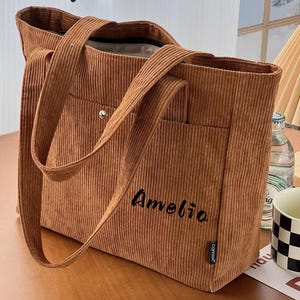 Può includere: Una borsa tote in velluto a coste marrone con il nome "Amelia" ricamato in nero. La borsa ha due manici, una tasca frontale e il testo "Gifts Best Ideas" in alto. La borsa è su una superficie di legno.
