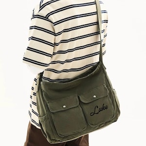 May include: An olive green canvas messenger bag with two front pockets and the name "Luke" embroidered on the front. The bag is worn over the shoulder. The person is wearing a striped shirt. The text "Gifts Best Ideas" is at the top.