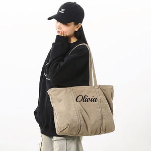 May include: A beige tote bag with the name "Olivia" printed on it. The bag is carried by a person wearing a black cap and a black sweatshirt. The text "Gifts Best Ideas" is at the top of the image.