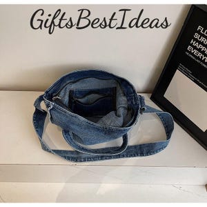 May include: A blue denim handbag with a shoulder strap. The bag is open, revealing an interior pocket with a zipper. The text "Gifts Best Ideas" is above the bag. A framed poster is in the background.