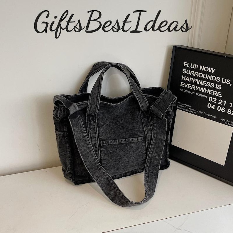 May include: A black denim tote bag with a shoulder strap and two handles. The bag has a front pocket and side pockets. The text "Gifts Best Ideas" is above the bag. A framed sign with text is to the right.