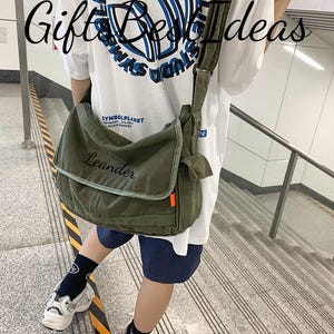 May include: An olive green messenger bag with the name "Leander" embroidered on the front flap. The bag has a long adjustable strap and a small side pocket with an orange accent. The background includes a white t-shirt and a staircase.