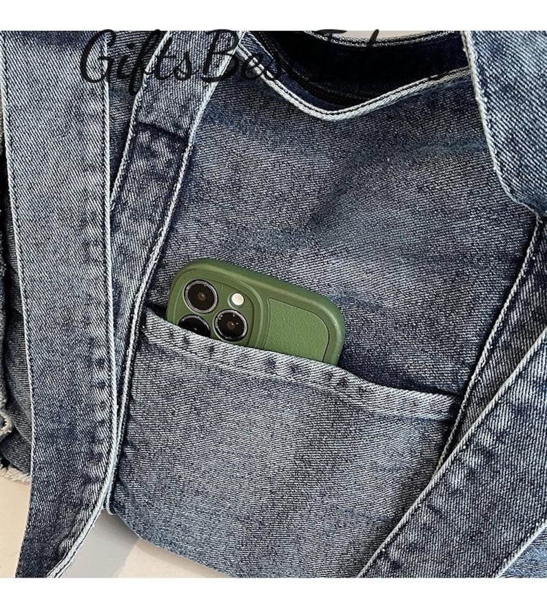 May include: Close-up of a denim tote bag with a pocket. The bag is a medium wash blue with white stitching and straps. A green phone case is visible in the pocket. The text "GiftsBox" is visible at the top.