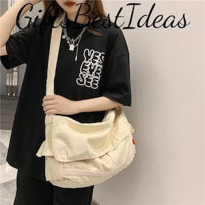 May include: A cream-coloured messenger bag with a large flap closure and a shoulder strap. The bag is worn over a black t-shirt with the text "YES EYE SEE" in white. The bag has a small red tag on the side.