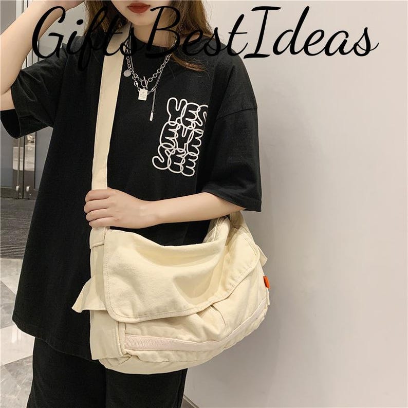 May include: A cream-coloured messenger bag with a large flap closure and a shoulder strap. The bag is worn over a black t-shirt with the text "YES EYE SEE" in white. The bag has a small red tag on the side.