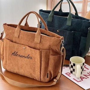 Peut inclure: Deux sacs fourre-tout en velours côtelé sont présentés. L'un est de couleur beige avec le nom "Alexander" brodé sur le devant. L'autre est bleu canard. Les deux ont des poignées et des bandoulières. Une tasse à motif damier noir et blanc est sur la table.