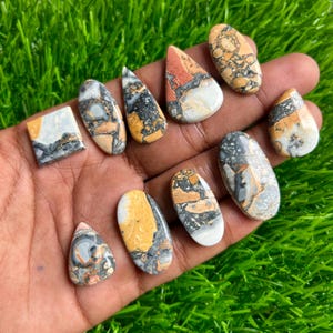May include: A collection of polished gemstones in various shapes, including oval, teardrop, and rectangular. The stones feature a mix of colors, such as yellow, orange, gray, black, and white, with unique patterns and textures. The gemstones are displayed on a green surface.