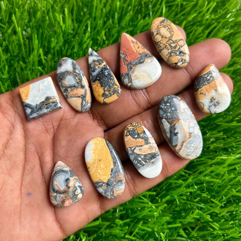 May include: A collection of polished gemstones in various shapes, including oval, teardrop, and rectangular. The stones feature a mix of colors, such as yellow, orange, gray, black, and white, with unique patterns and textures. The gemstones are displayed on a green surface.
