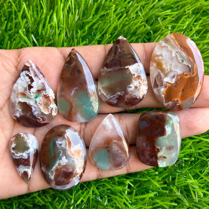 May include: A collection of polished gemstones in various shapes, including teardrop and oval. The stones display a mix of colors, including brown, white, and teal, with unique patterns and textures. The gemstones are displayed on a bed of green grass.