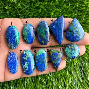 May include: Ten azurite gemstones in diverse shapes, including ovals and triangles, are shown. The stones exhibit a blend of deep blue and green colours, with distinctive patterns. The gemstones are displayed on a hand against a green background.