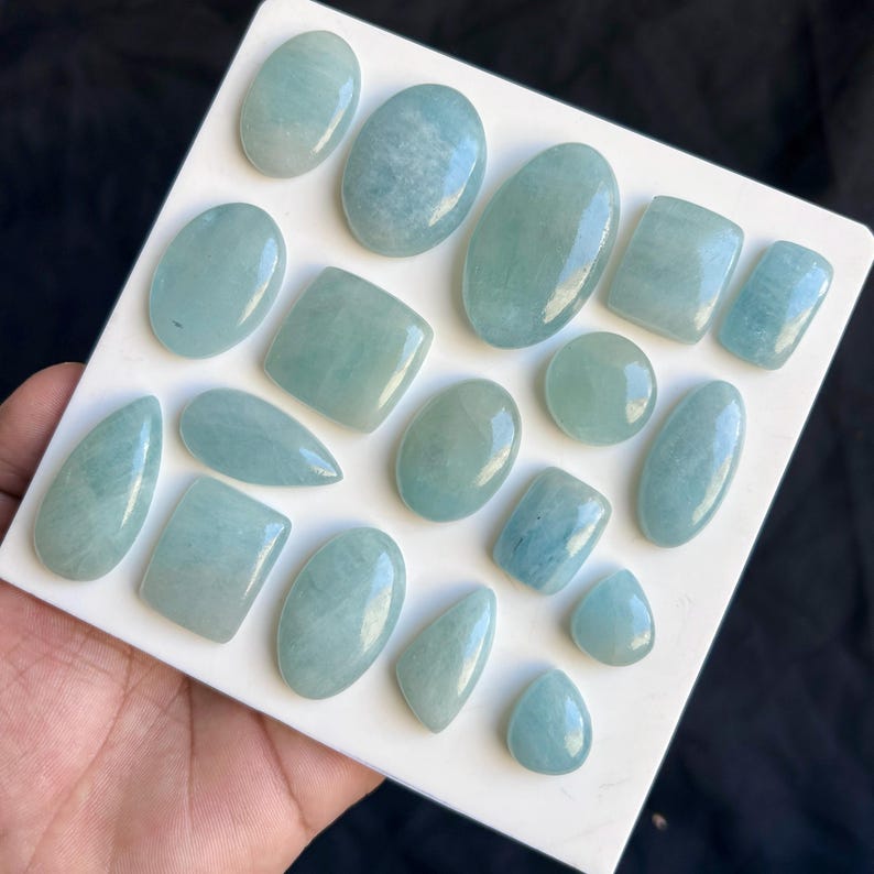 May include: A selection of aquamarine gemstones in diverse forms, such as ovals, squares, and teardrops. The gemstones exhibit a pale blue-green hue and are presented on a white surface. The polished gemstones have a smooth, reflective finish.