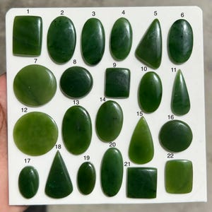 Op de afbeelding: Een verzameling van tweeëntwintig gepolijste groene jade cabochons gerangschikt op een wit displaybord. Elke steen is genummerd van 1 tot 22 en heeft verschillende vormen, waaronder ovalen, cirkels, vierkanten en driehoeken met gladde oppervlakken.