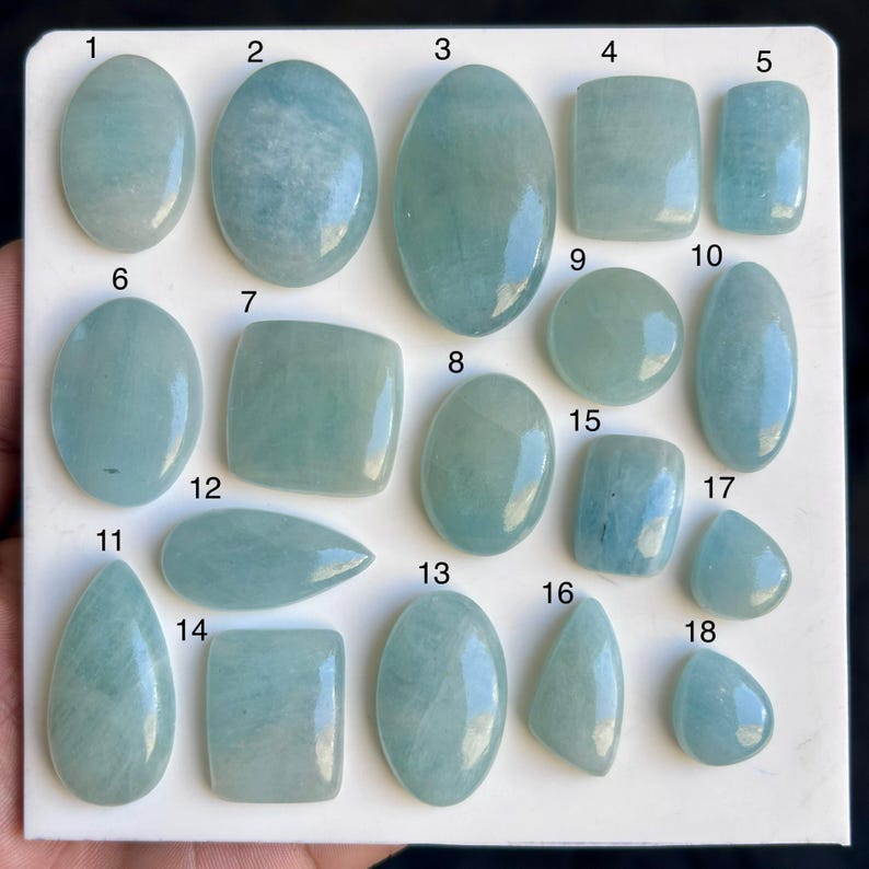 May include: A selection of aquamarine gemstones in diverse forms, such as oval, rectangular, and teardrop. The gemstones exhibit a pale blue-green hue and are displayed on a white surface. Each stone is numbered for easy reference.