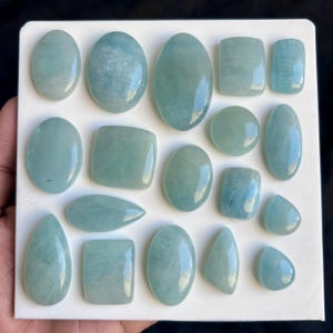 May include: A selection of aquamarine gemstones in diverse shapes, such as ovals, rectangles, and teardrops. The gemstones exhibit a pale blue-green hue and are arranged on a white surface. The polished gemstones have a smooth, reflective appearance.