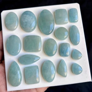 May include: A selection of aquamarine gemstones in diverse forms, such as oval, rectangular, and teardrop. The gemstones exhibit a pale blue-green hue and are presented on a white surface. The stones are polished, displaying a smooth, reflective finish.