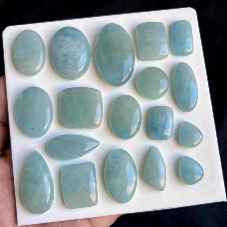 May include: A selection of aquamarine gemstones in diverse forms, such as oval, rectangular, and teardrop. The gemstones exhibit a pale blue-green hue and are presented on a white surface. The stones are polished, displaying a smooth, reflective finish.