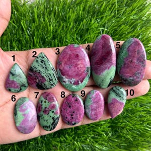 Puede incluir: Una colección de diez piedras preciosas pulidas, cada una con una forma y un patrón únicos. Las piedras presentan una mezcla de colores, incluyendo rojo intenso, verde y negro. Las piedras preciosas se sostienen en una mano sobre un fondo de hierba verde.