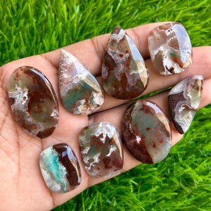 May include: A collection of eight polished gemstones in various shapes, including oval, teardrop, and rectangular. The stones display a mix of colors, including brown, green, and white, with unique patterns and textures. The gemstones are displayed on a hand.
