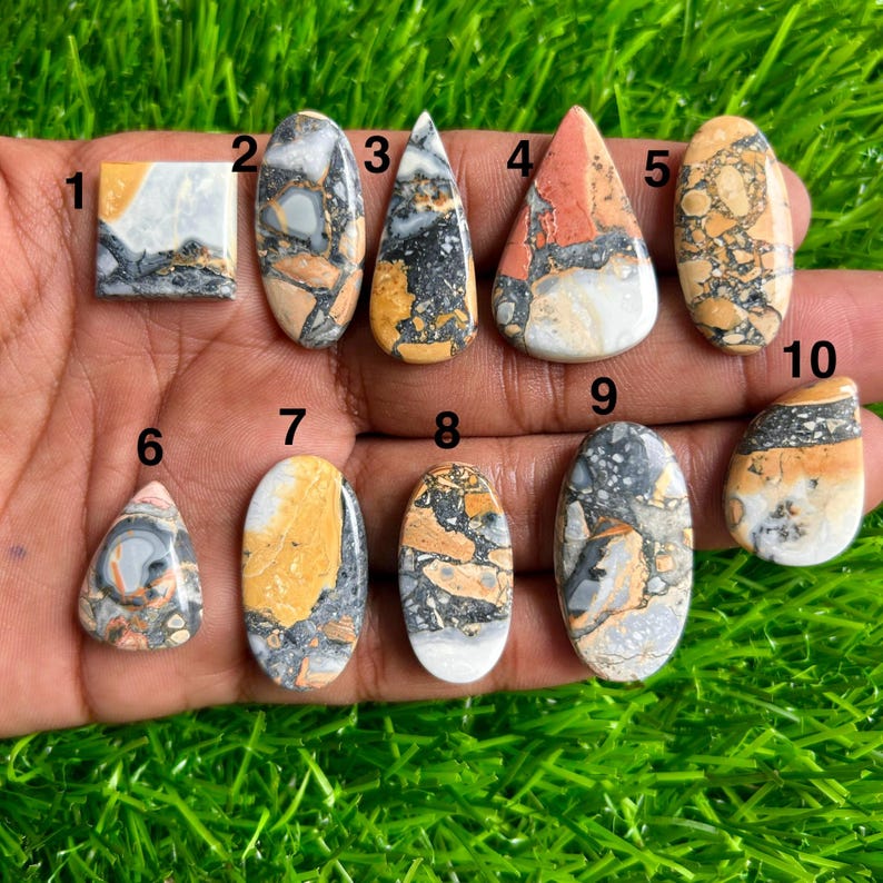 May include: A collection of ten polished gemstones in various shapes, including square, oval, and teardrop. The stones display a mix of colors, such as beige, gray, black, and orange, with unique patterns and textures. The gemstones are displayed on a bed of green grass.