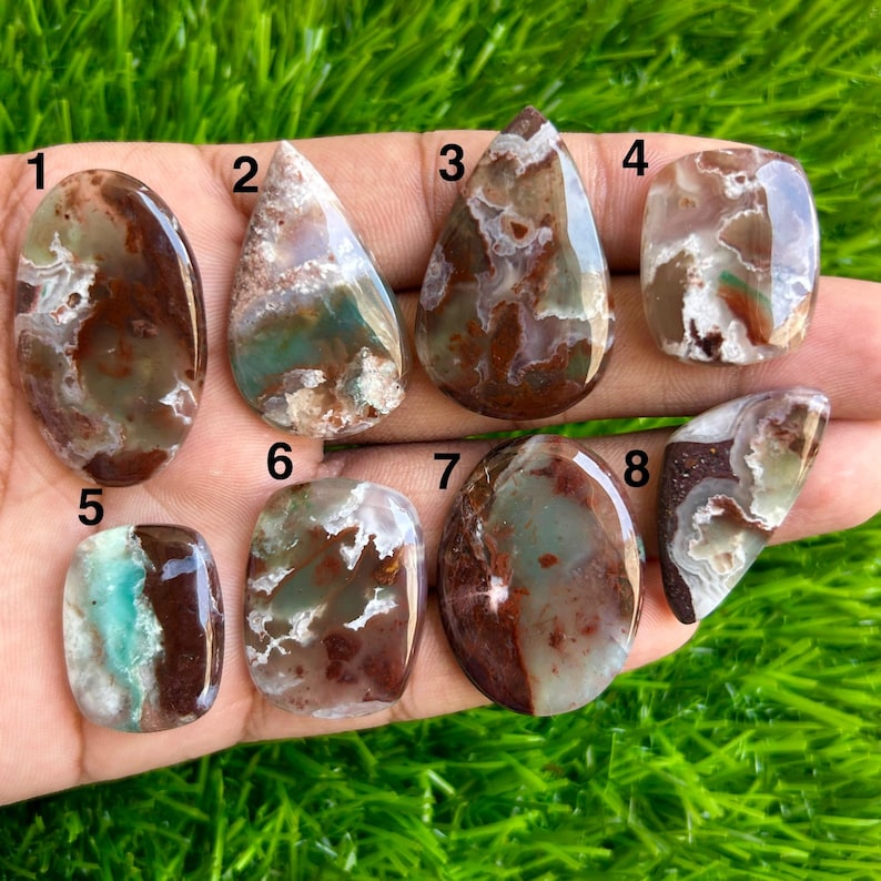 May include: A collection of eight polished gemstones in various shapes, including oval, teardrop, and rectangular. The stones feature a mix of brown, green, and white colors, with unique patterns and textures. The gemstones are displayed against a green background.