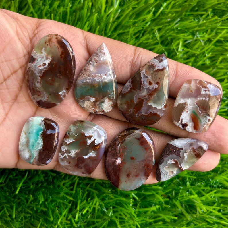 May include: A collection of eight polished gemstones in various shapes, including oval and teardrop. The stones feature a mix of brown, white, and teal colors, with unique patterns and textures. The gemstones are displayed on a hand against a backdrop of green grass.