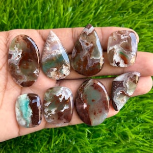 May include: A collection of polished gemstones in various shapes, including oval, teardrop, and rectangular. The stones feature a mix of brown, green, and white colors, with unique patterns and textures. The gemstones are displayed against a green background.