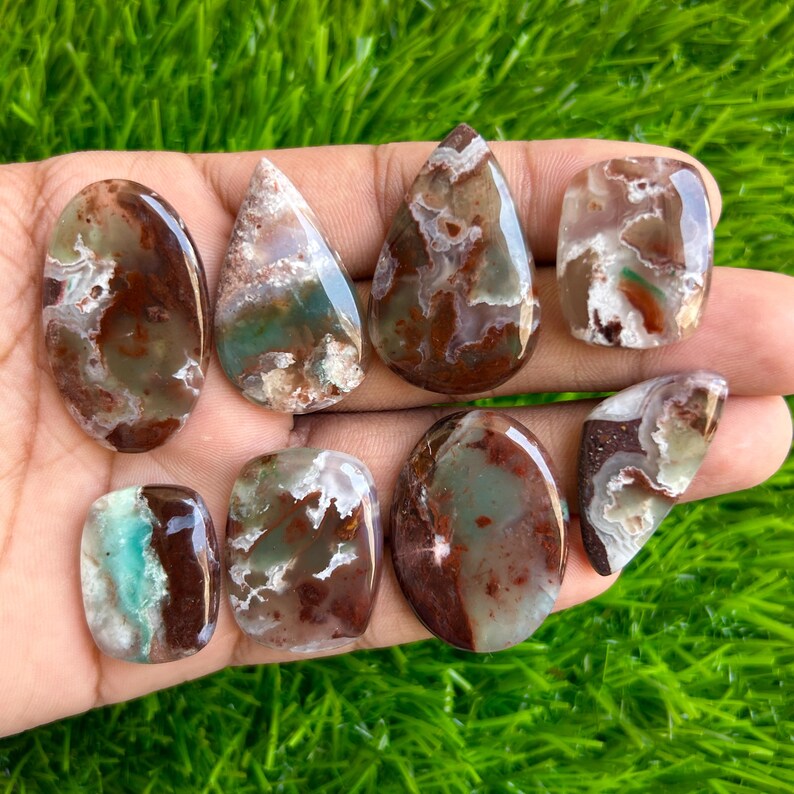 May include: A collection of polished gemstones in various shapes, including oval, teardrop, and rectangular. The stones feature a mix of brown, green, and white colors, with unique patterns and textures. The gemstones are displayed against a green background.