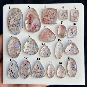 May include: Nineteen polished gemstones of different shapes, including oval, teardrop, and square. The translucent stones have red and black inclusions within a clear or tinted base. The gemstones are displayed on a white surface. Measurements are in cm.