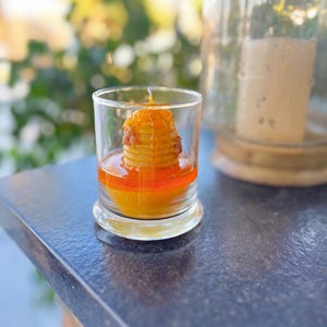May include: A clear glass candle holder with a stack of orange-colored wax, resembling pancakes, and a wick. The candle sits in a pool of orange wax. A glass cloche is in the background.
