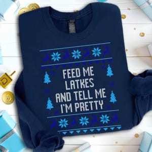 Może przedstawiać: Granatowa bluza z świątecznym wzorem. Tekst "FEED ME LATKES AND TELL ME I'M PRETTY" jest wyświetlany na biało. Wzór zawiera płatki śniegu, renifery i niebieskie choinki.