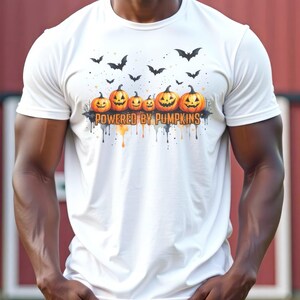 May include: White t-shirt featuring a row of carved orange pumpkins with smiling faces, and black bats flying above. The text "POWERED BY PUMPKINS" is printed below the pumpkins. The design has orange and black watercolor drips.