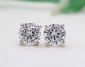 May include: A pair of round-cut diamond stud earrings. The diamonds are set in a silver-coloured metal. The earrings are displayed on a white surface with a blurred green and white background. The Aurista Jewels logo is visible in the upper right corner.