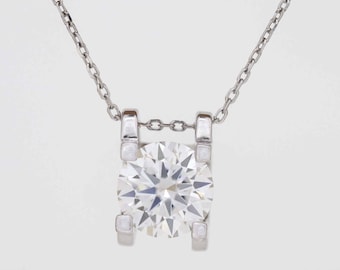 Puede incluir: Un collar de plata con un colgante de diamante redondo y brillante. El diamante está sujeto por un engaste de cuatro garras, suspendido de una delicada cadena. El colgante y la cadena son de metal plateado brillante.