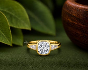 May include: A gold ring featuring a central cushion-cut stone in a bezel setting, flanked by two smaller triangular-cut stones on each side. The band is polished gold, and the text Aurista Jewelry is visible near the central setting.