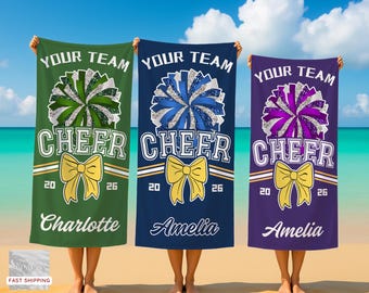 Peut inclure: Trois serviettes de cheerleading personnalisées, vertes, bleues et violettes, chacune avec un motif de pompon et le texte "YOUR TEAM CHEER". Les serviettes comportent également un nœud, les années "20 26" et un nom.