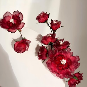 Puede incluir: Un grupo de flores de tela rojo oscuro con centros blancos. Las flores están dispuestas sobre una superficie blanca, con algunas flores completamente abiertas y otras en forma de capullo. Los pétalos tienen una calidad delicada y transparente.