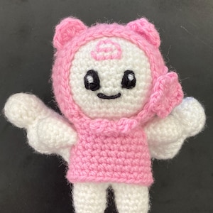 May include: A handmade crochet doll in white and pink. The doll has a pink hood with bear ears, black eyes, and a small smile. It is wearing a pink dress and has white wings.